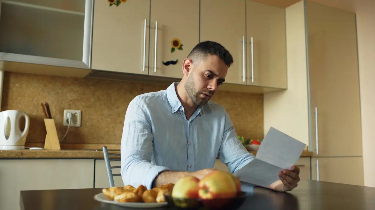 Homem sentado à mesa da cozinha lendo uma carta ao lado de uma xícara de café