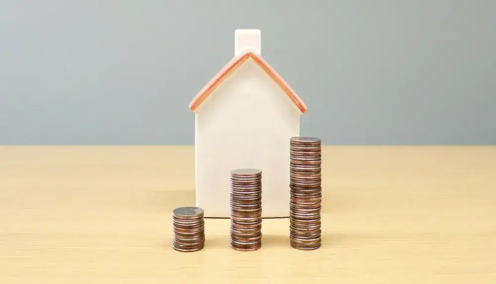 Miniatura de casa branca ao lado de pilhas de moedas sobre mesa de madeira