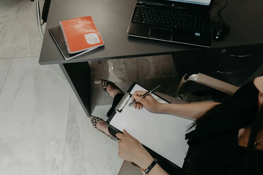 Pessoa sentada à mesa conferindo papéis ao lado de um notebook