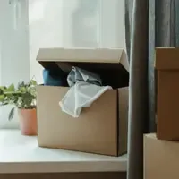 Caixa de papelão aberta, com roupas saindo pela tampa, sobre o parapeito de uma janela, ao lado de um vaso de planta e de outras caixas fechadas